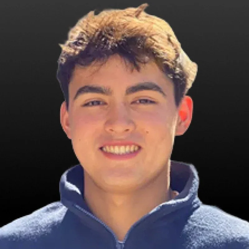 A young man smiling outdoors with a dark background, wearing a blue jacket.