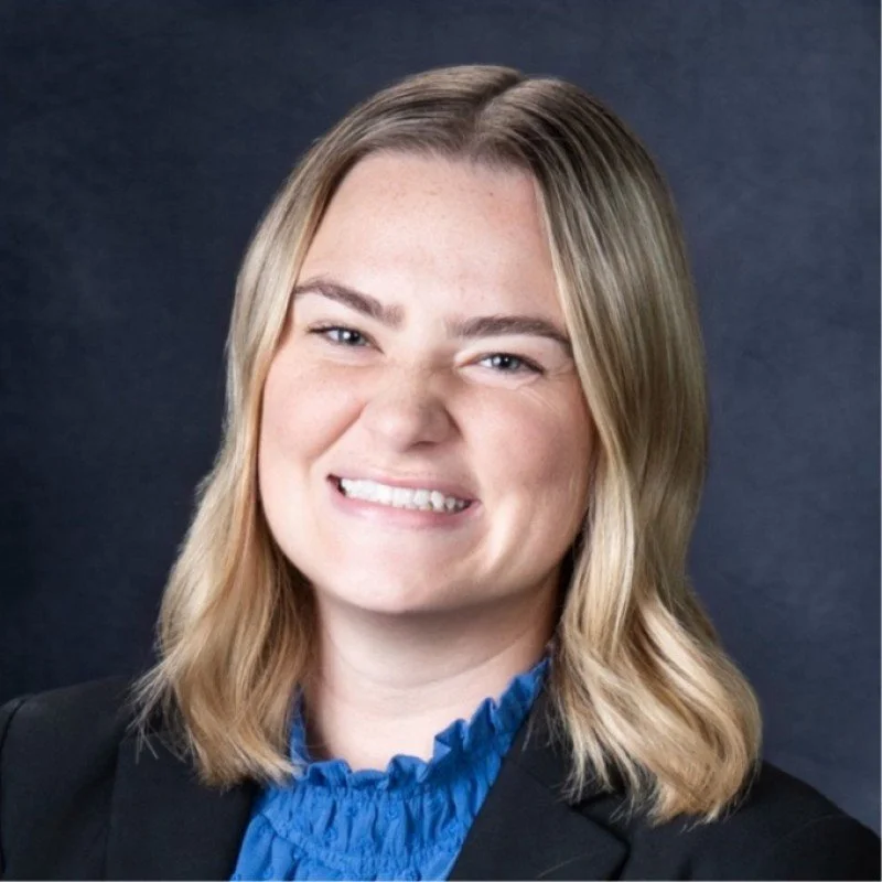 A professional woman with shoulder-length blonde hair, smiling with a slight curl, wearing a black blazer over a blue ruffled blouse, against a dark grey background.