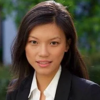 A woman with straight dark hair, wearing a white shirt and black blazer, smiling outdoors.
