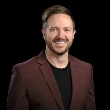 A man with short brown hair and a beard, wearing a maroon blazer and black shirt, smiling against a black background.