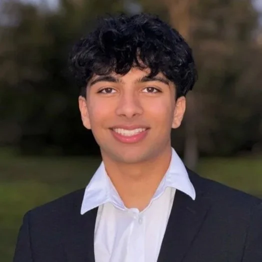 Portrait of a young man with dark, curly hair smiling outdoors, wearing a navy blazer and white shirt.