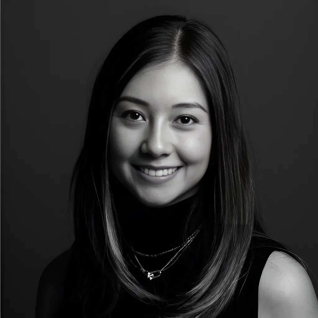 A black and white portrait of a young woman with straight hair, smiling, wearing a dark sleeveless top and layered necklaces, against a dark background.