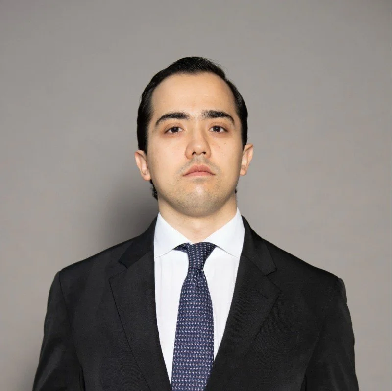 A young man in a black suit, white shirt, and patterned tie, standing against a plain gray background.