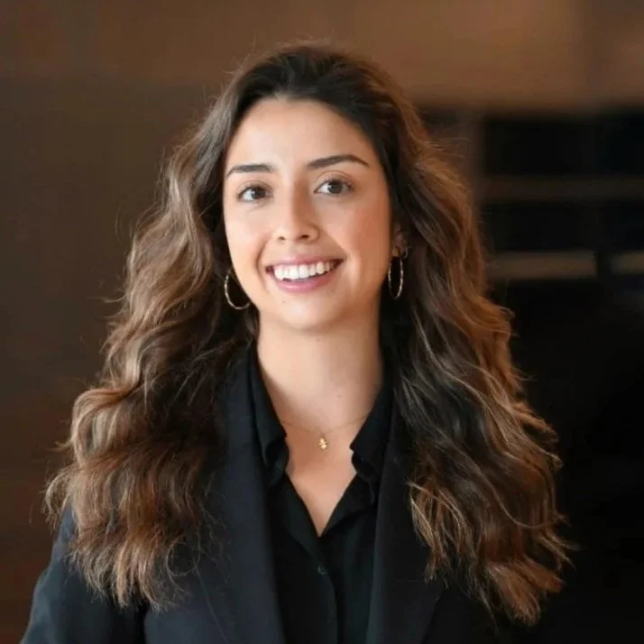 A young woman with long, wavy brown hair smiling at the camera, wearing a black blazer, gold hoop earrings, and a delicate gold necklace, in an indoor setting with a blurred background.