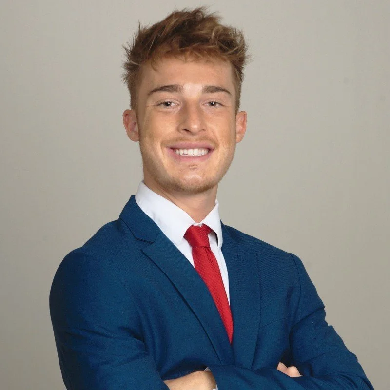 A young man in a blue suit, white shirt, and red tie standing against a neutral background, smiling confidently with arms crossed.