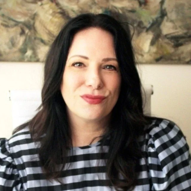 A woman with dark hair and red lipstick smiling, wearing a black and white checkered shirt, sitting in front of a wall with abstract art.