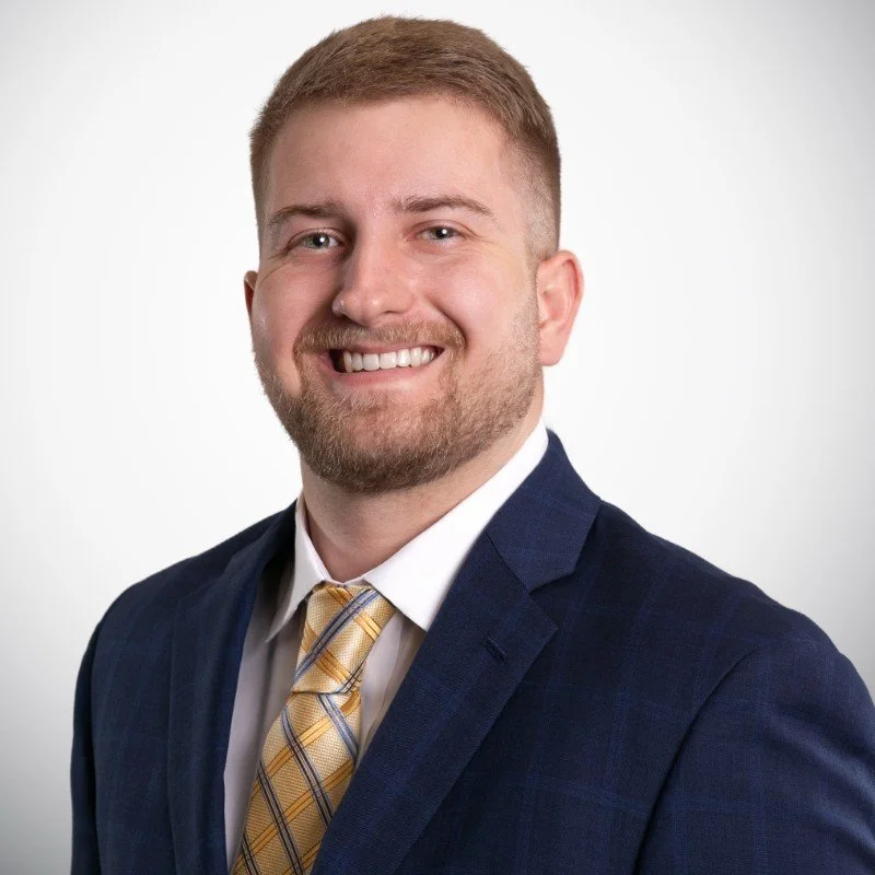 A portrait of a smiling man with short light brown hair and a beard, wearing a navy blazer, white shirt, and yellow plaid tie, against a plain white background.