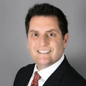 Professional headshot of a man with dark hair, smiling, wearing a dark pinstripe suit and white shirt against a gray background.