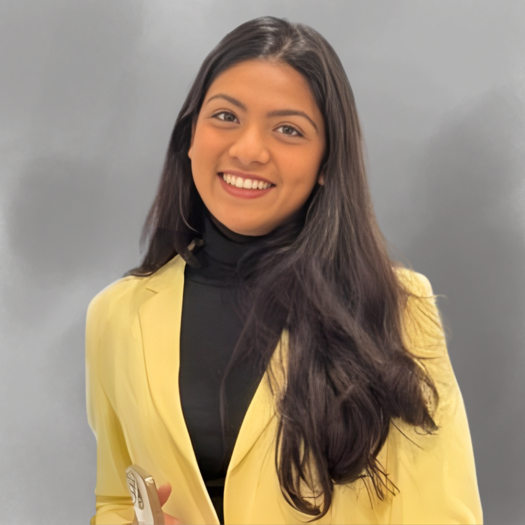 Young woman smiling, wearing a yellow blazer over a black turtleneck, with long dark hair, posing against a gray background.