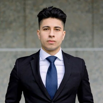 A young man in a black suit, white shirt, and blue tie standing outdoors against a concrete wall.