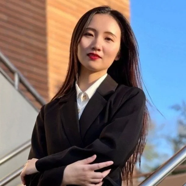 Young woman with long dark hair wearing a black blazer and white shirt, standing outdoors near stairs, with a modern building and clear sky in the background.