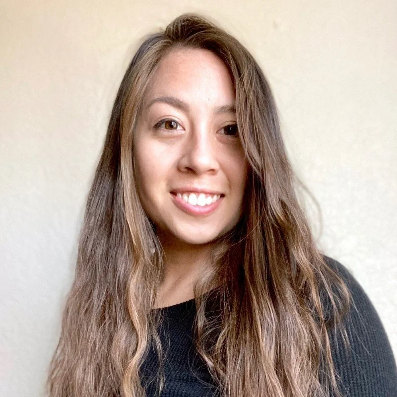 A young woman with long wavy brown hair smiling, wearing a black shirt, indoors with a plain light-colored wall background.