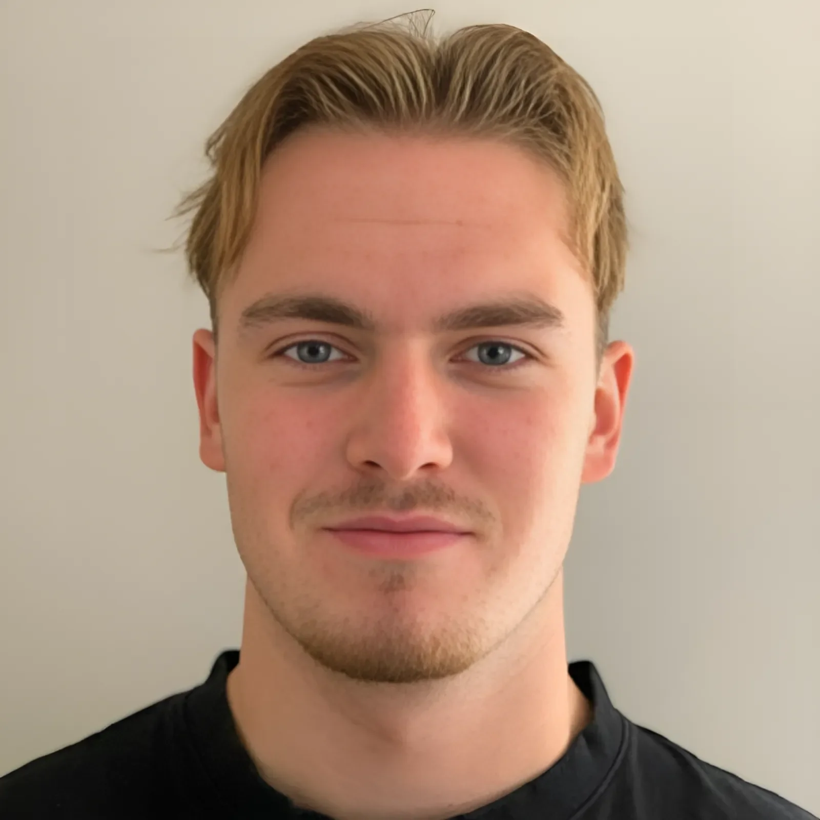 Close-up of a young man with short, light brown hair, blue eyes, slight facial hair, and a black shirt, standing against a plain light-colored wall.