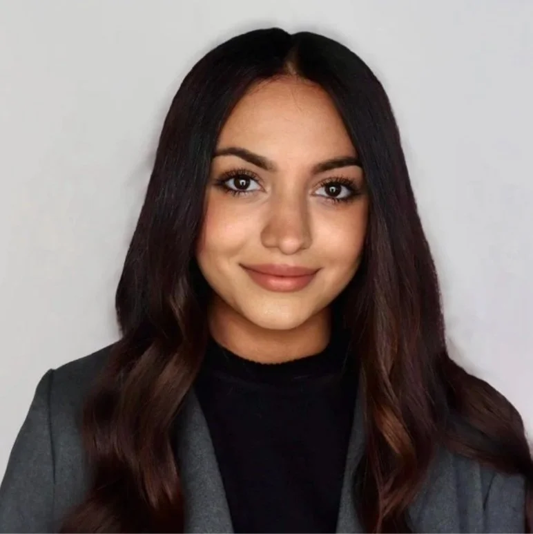Young woman with long dark hair, smiling, wearing a black top and gray blazer against a plain white background.