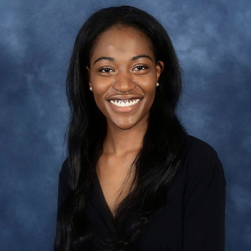 Headshot portrait of a young African American woman with long black hair, smiling, wearing a black top, against a blue mottled background.