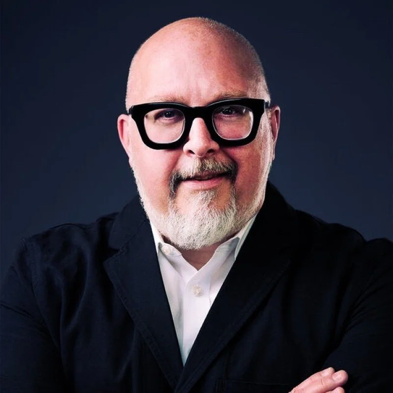 A middle-aged man with a bald head, glasses, and a white beard, wearing a black blazer and white shirt, is smiling and crossing his arms, against a dark background.