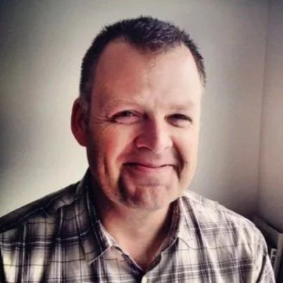 A smiling middle-aged man with short dark hair, wearing a plaid shirt, standing indoors against a plain wall.