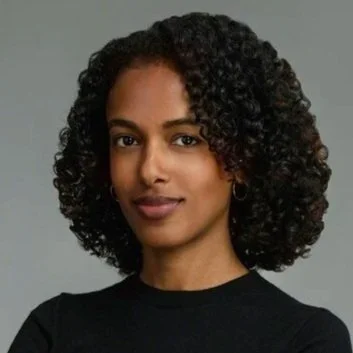 Portrait of a woman with curly black hair wearing a black top, looking at the camera against a neutral background.