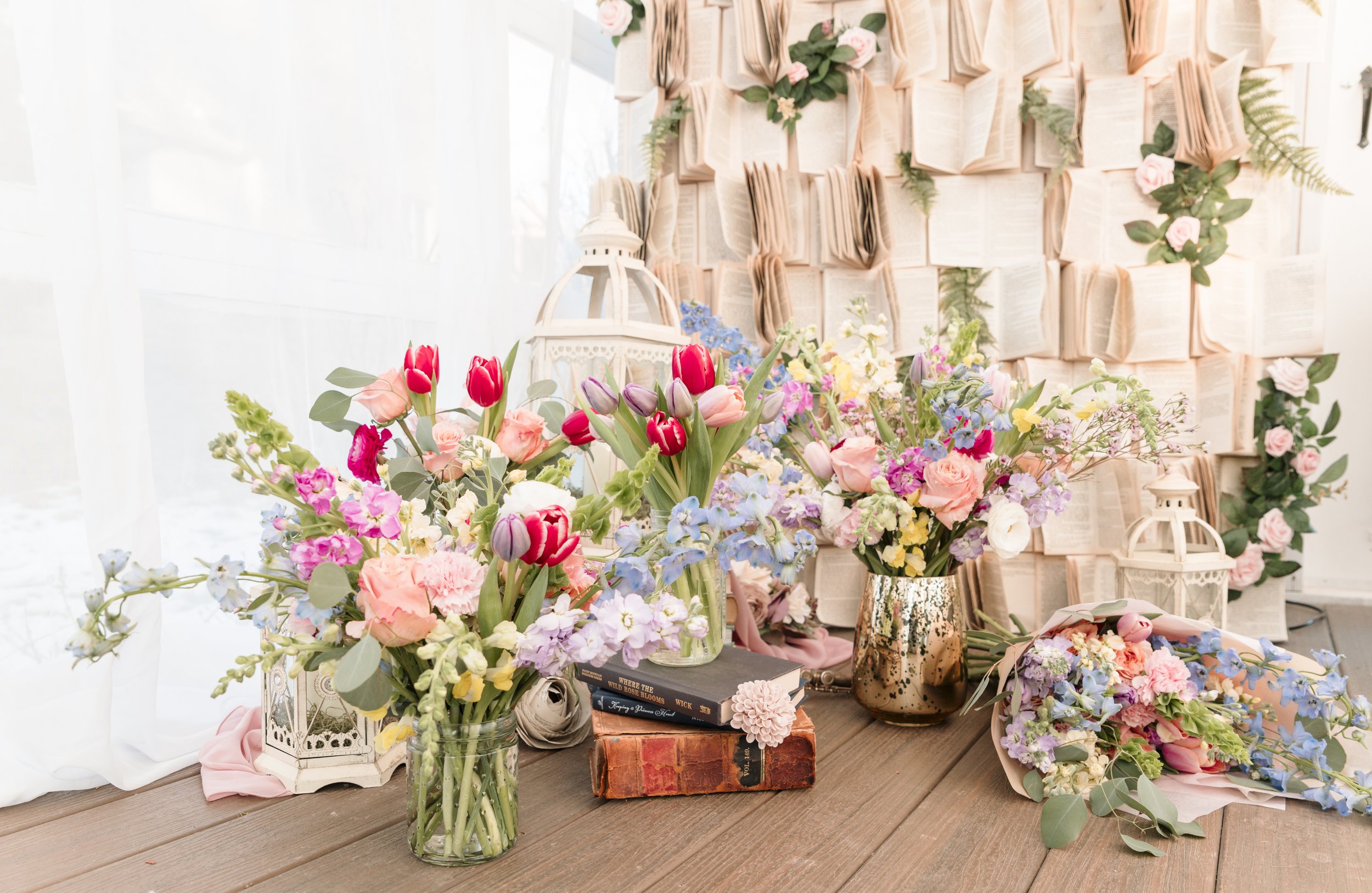 Arranged floral bouquets with pink, red, purple, and blue flowers on a wooden table, decorative books, lanterns, and a wall decorated with open books and pink roses in the background.