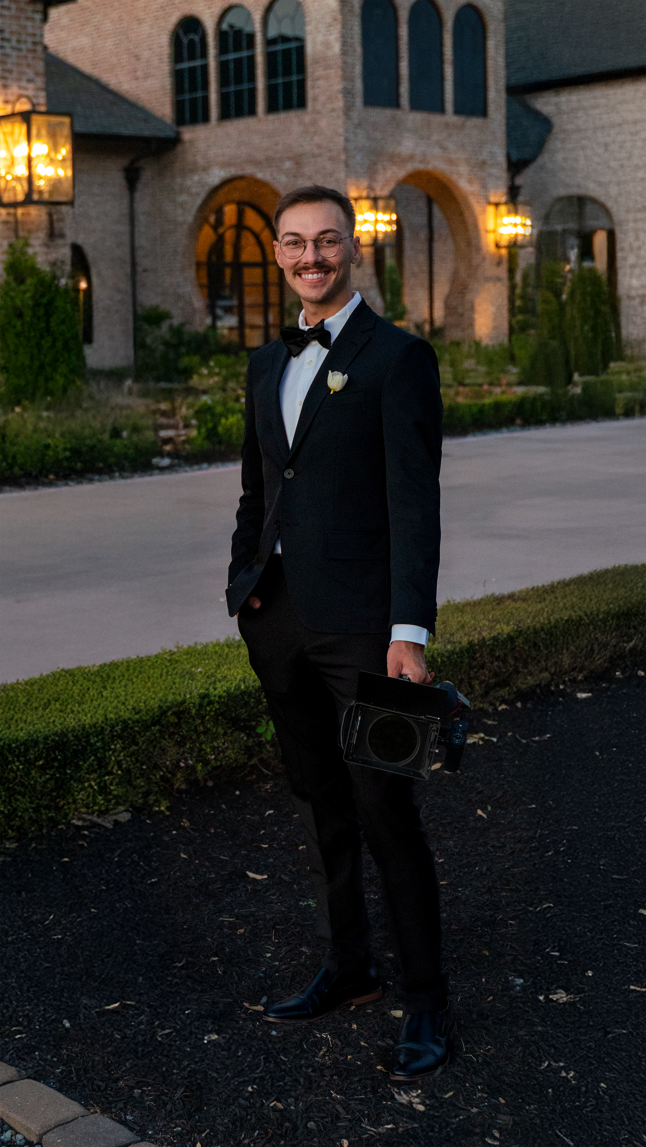 A young man in a tuxedo with a bow tie, holding a camera, stands outdoors in front of a large brick house with lit lanterns, during dusk.