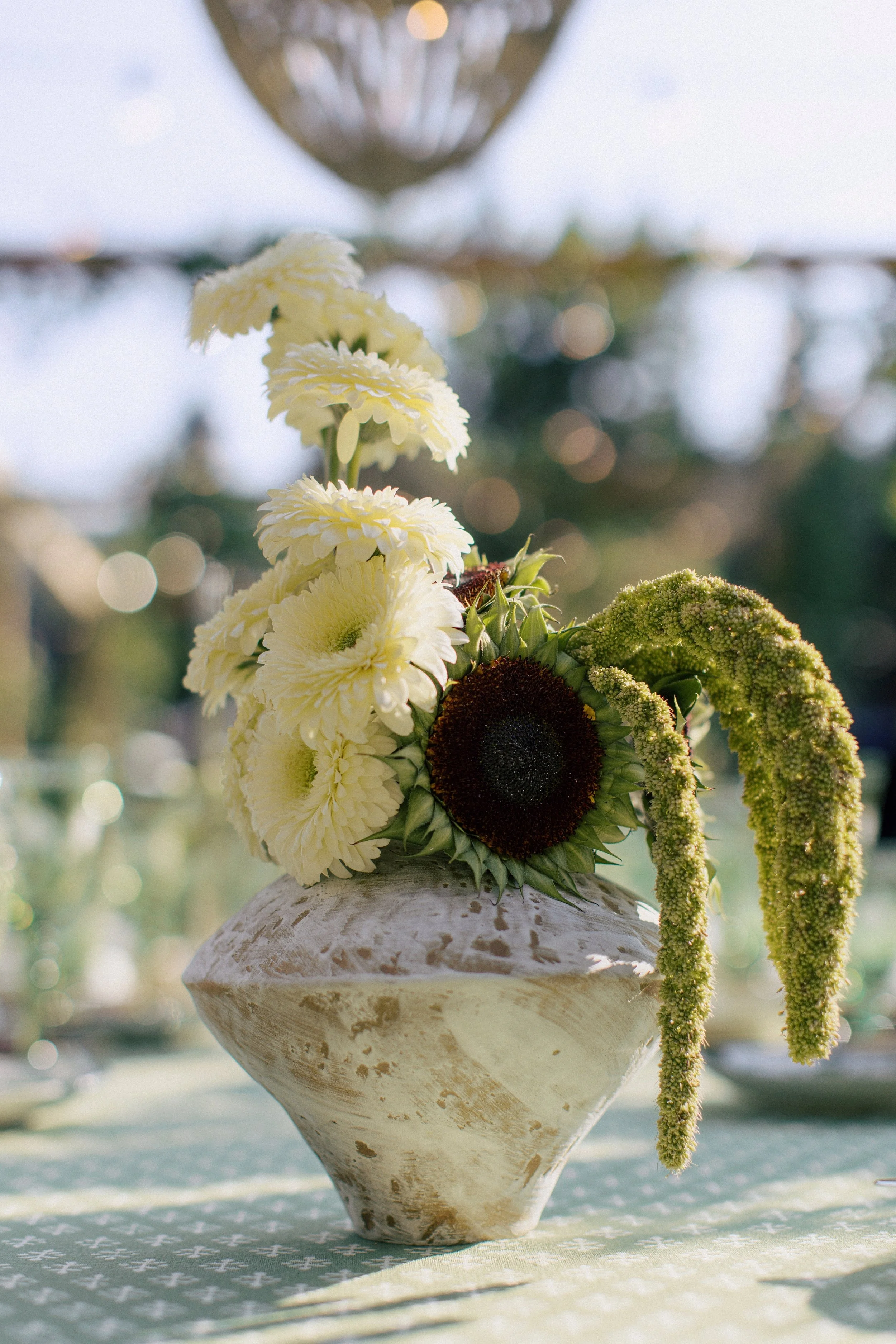A bouquet of white and green flowers, including sunflowers, arranged in a rustic white vase on a table with a patterned cloth, outdoors with sunlight.