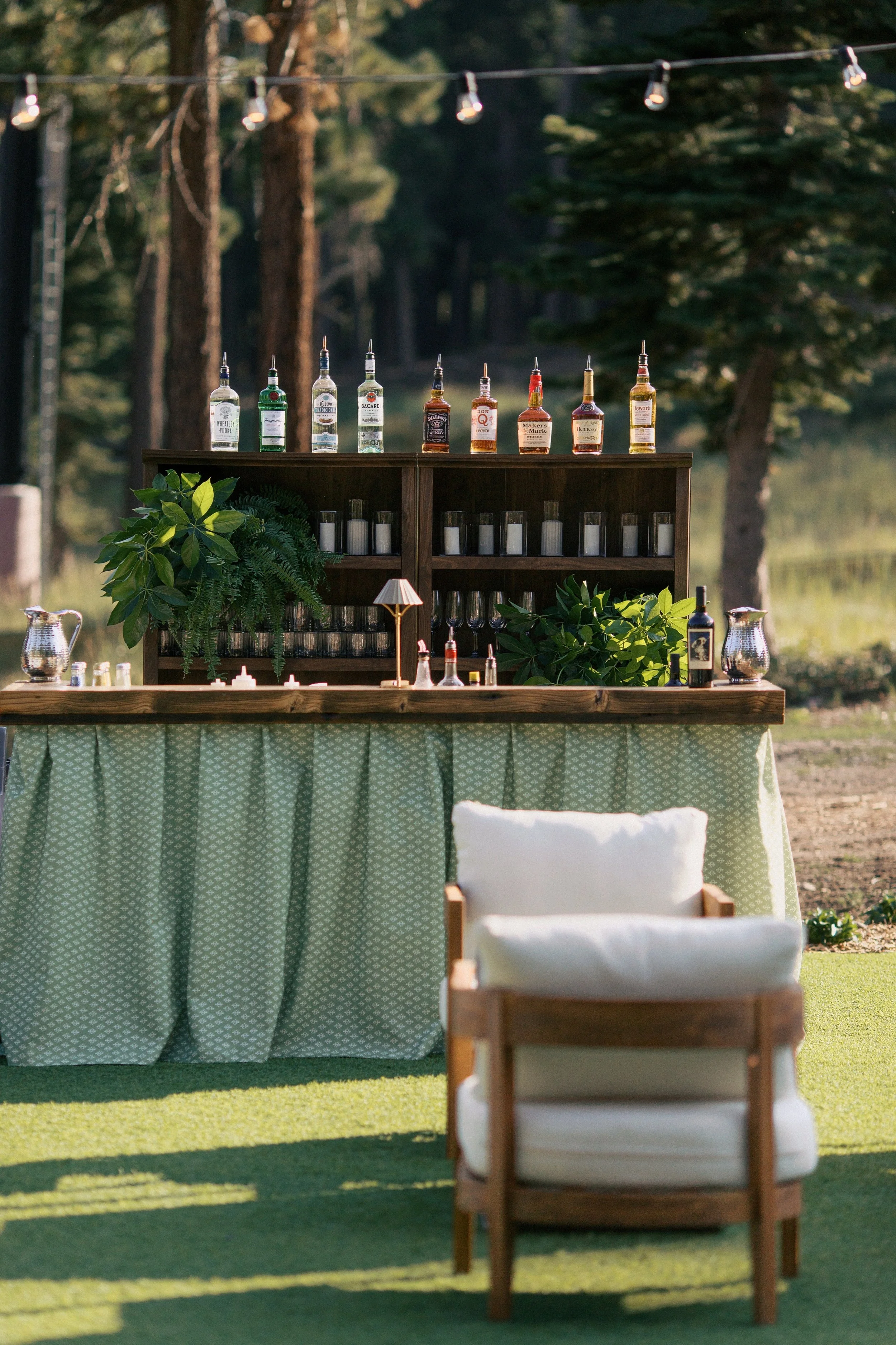 Outdoor bar setup with various liquor bottles, glassware, plants, and a small lamp, in a natural setting with trees and water in the background.