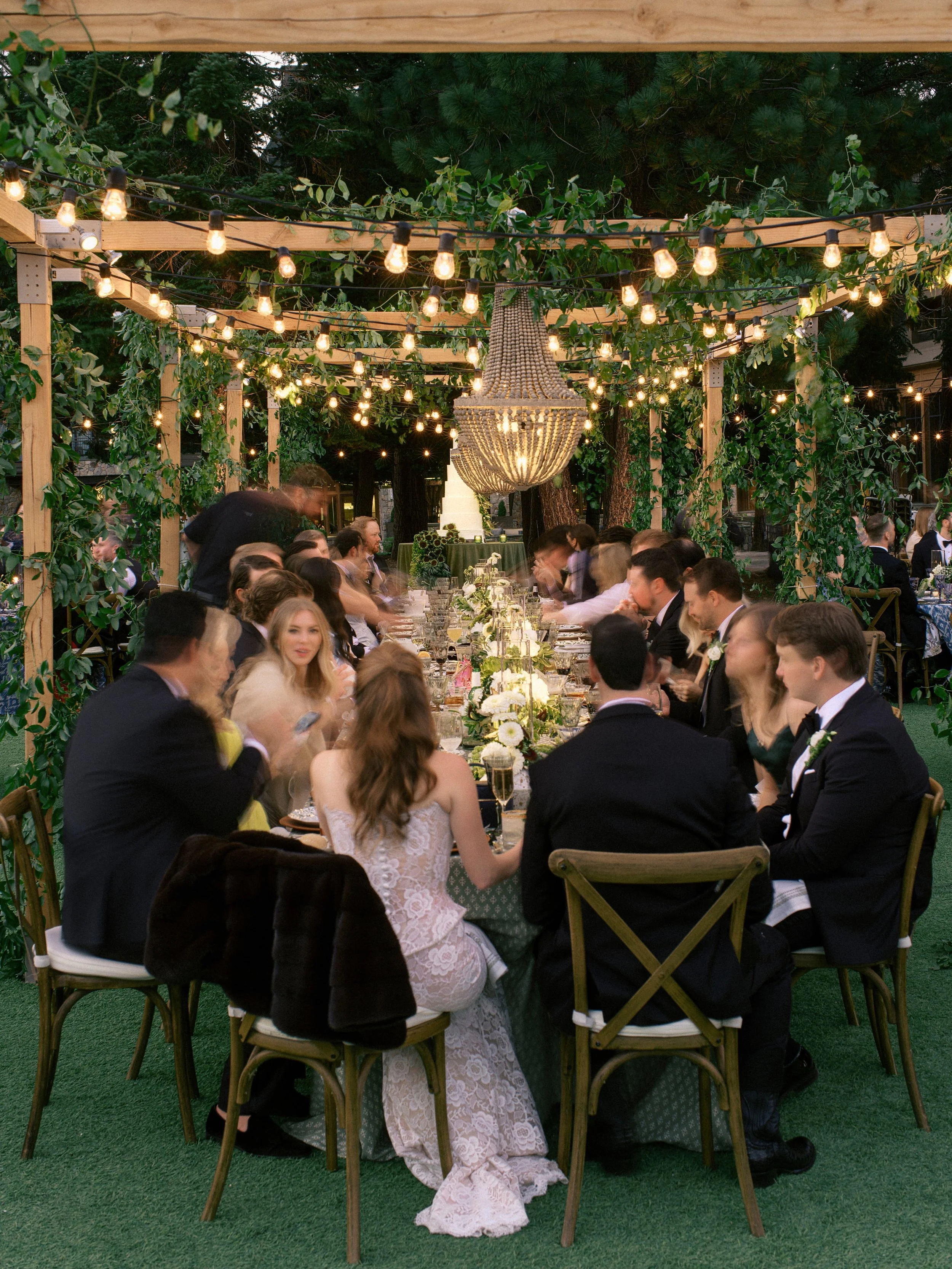 People dressed in formal attire sitting around a long outdoor banquet table decorated with flowers and candles, under string lights and a large chandelier hanging from wooden pergola, surrounded by trees.