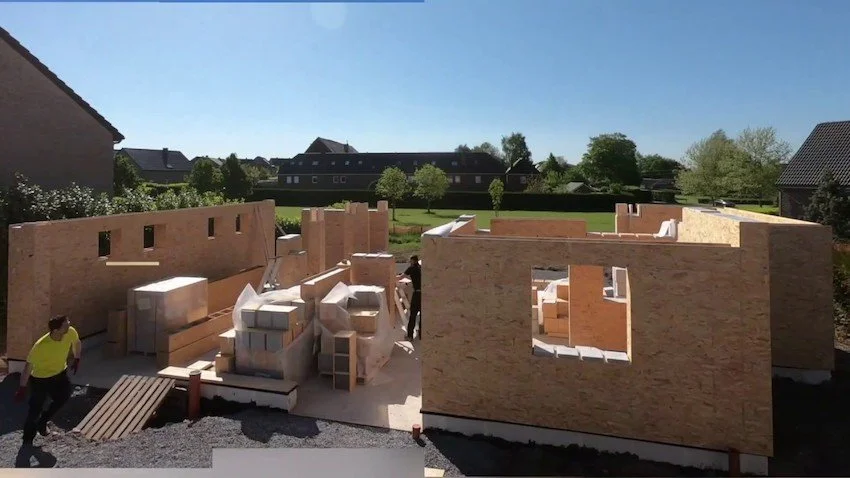 Construction site with wooden panels and partially built walls, workers in yellow shirts, residential neighborhood in the background, clear sky.