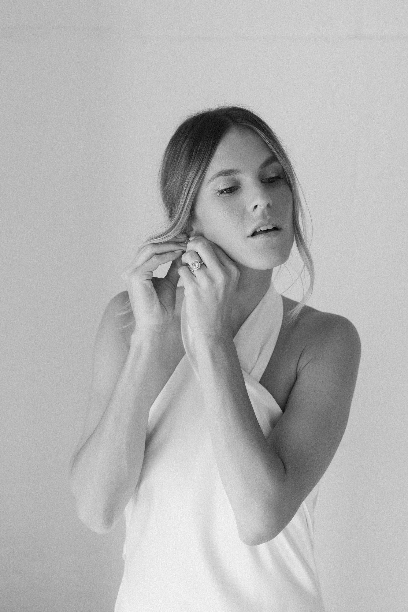  a modern black and white photo of the bride getting ready while putting in her pearl stud earrings by abellie wearing her silk wedding dress. 