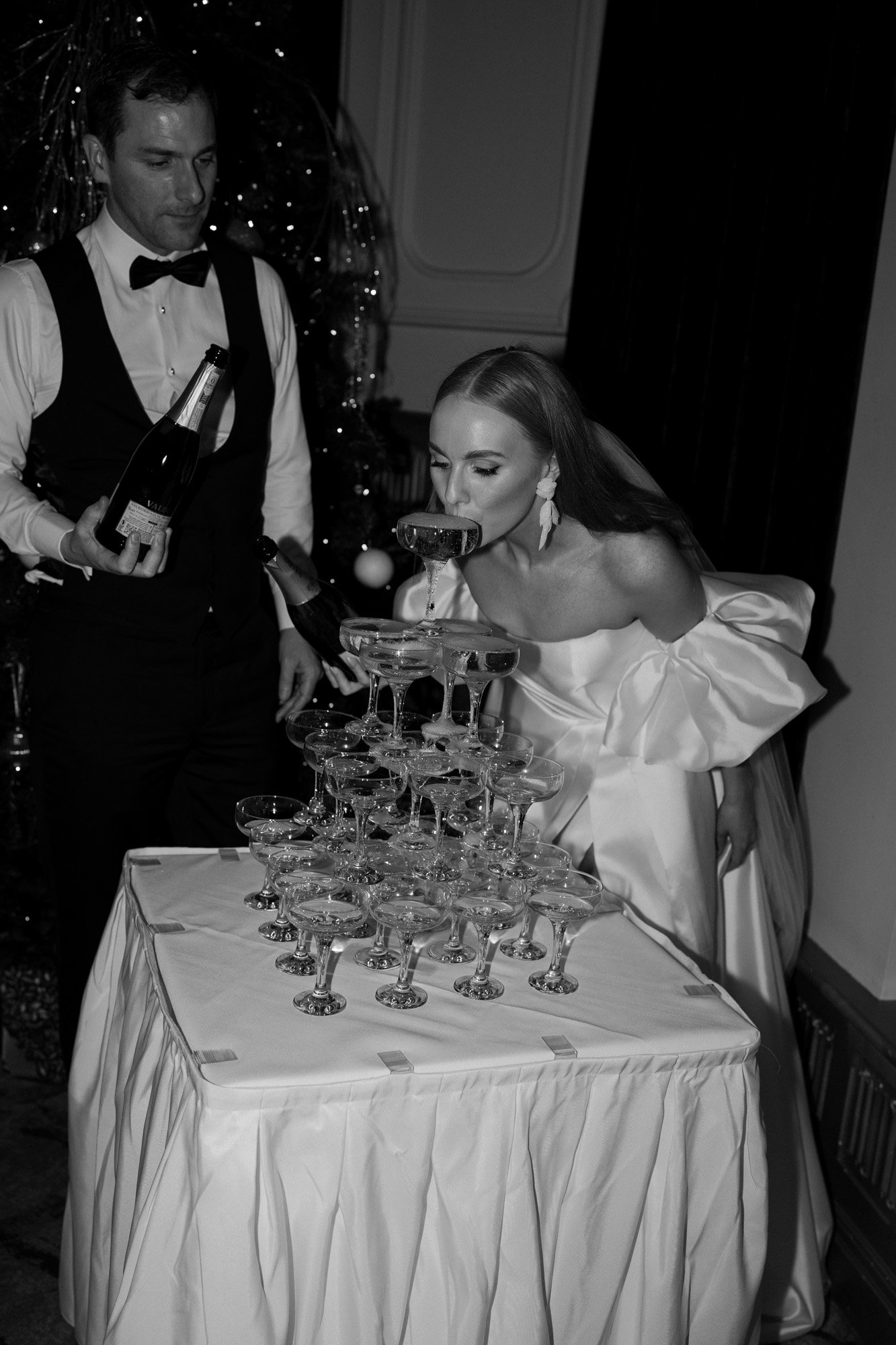 a modern champagne tower being poured by the groom as the bride sips playfully from a coupe wearing her strapless silk ball gown by alena leena and abellie earrings.