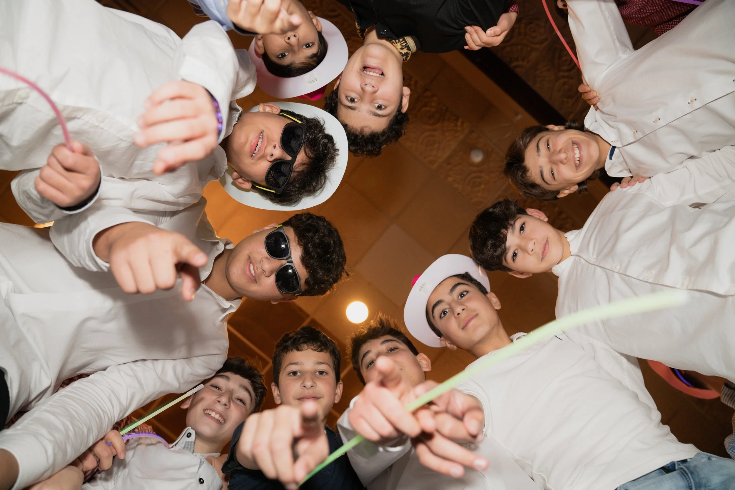A group of children wearing white clothes and hats, some wearing sunglasses, looking down at the camera and smiling.