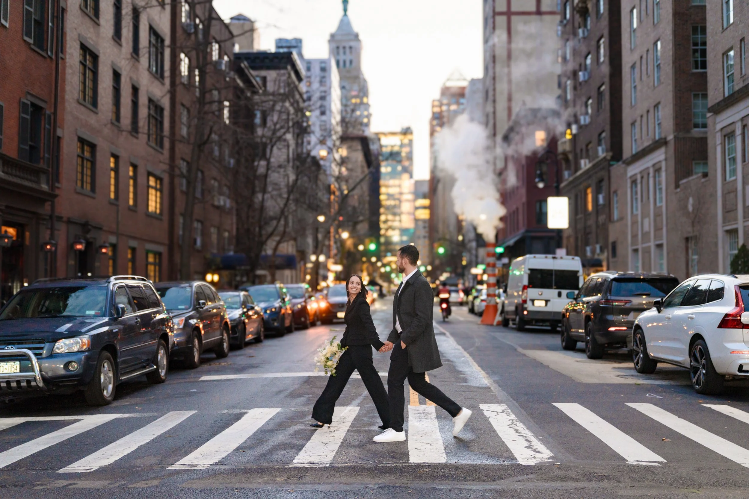 A couple dressed in formal attire holding hands and walking across a city street crosswalk with parked cars on both sides, tall brick buildings, and steam rising from a manhole in the background.