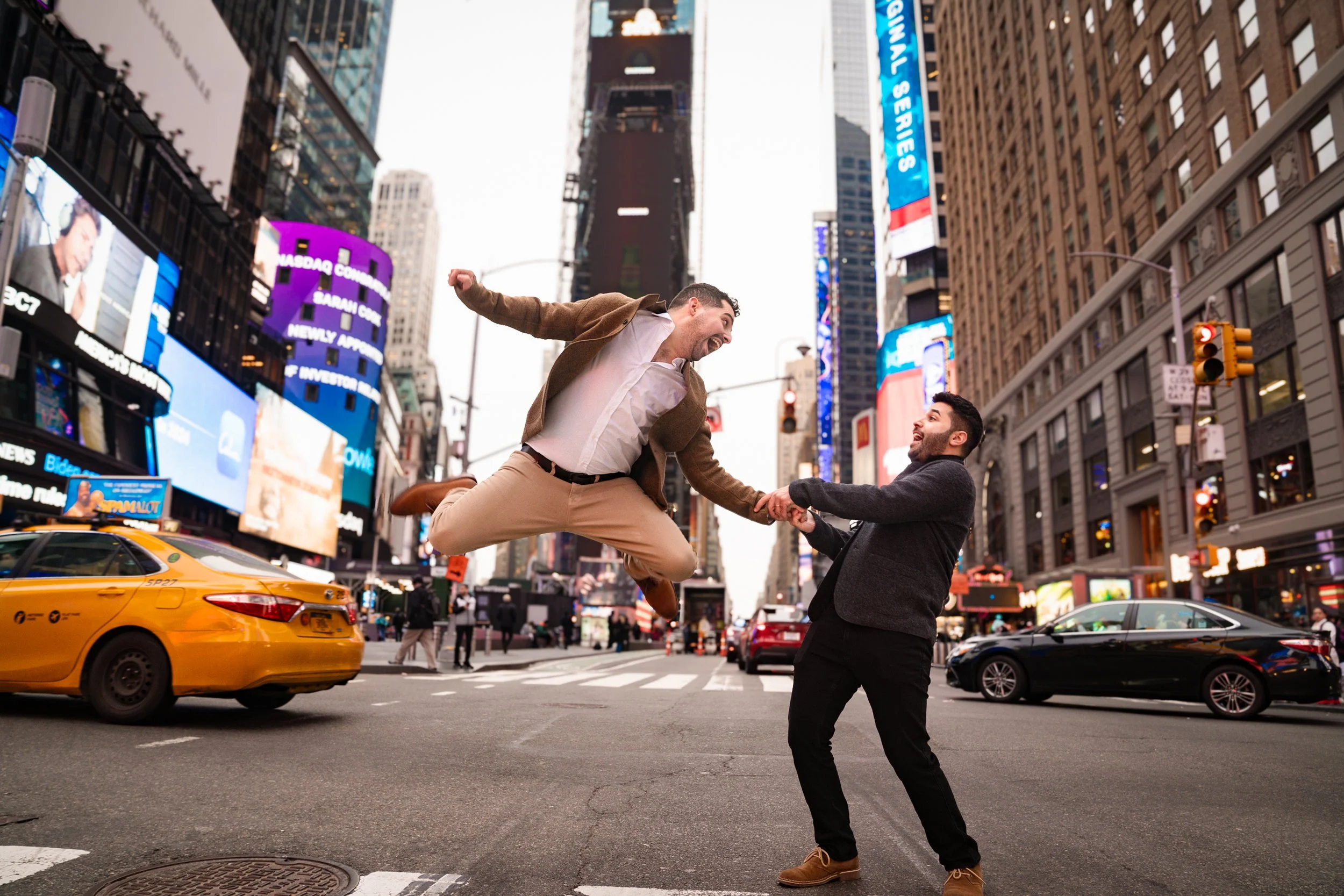 Two men in business casual attire are holding hands and dancing in the middle of a city street with tall buildings and large digital billboards, including a yellow taxi cab parked nearby, looking joyous and energetic.