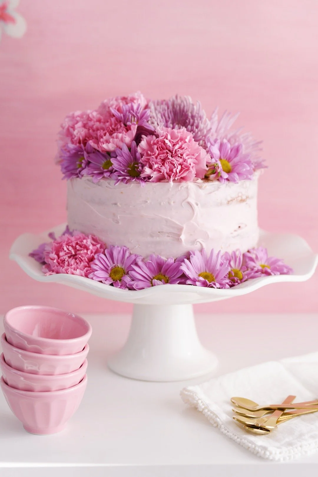 Pink frosted cake decorated with pink and purple flowers on a white cake stand, with pink bowls, gold utensils, and a white napkin next to it, all against a pink background.