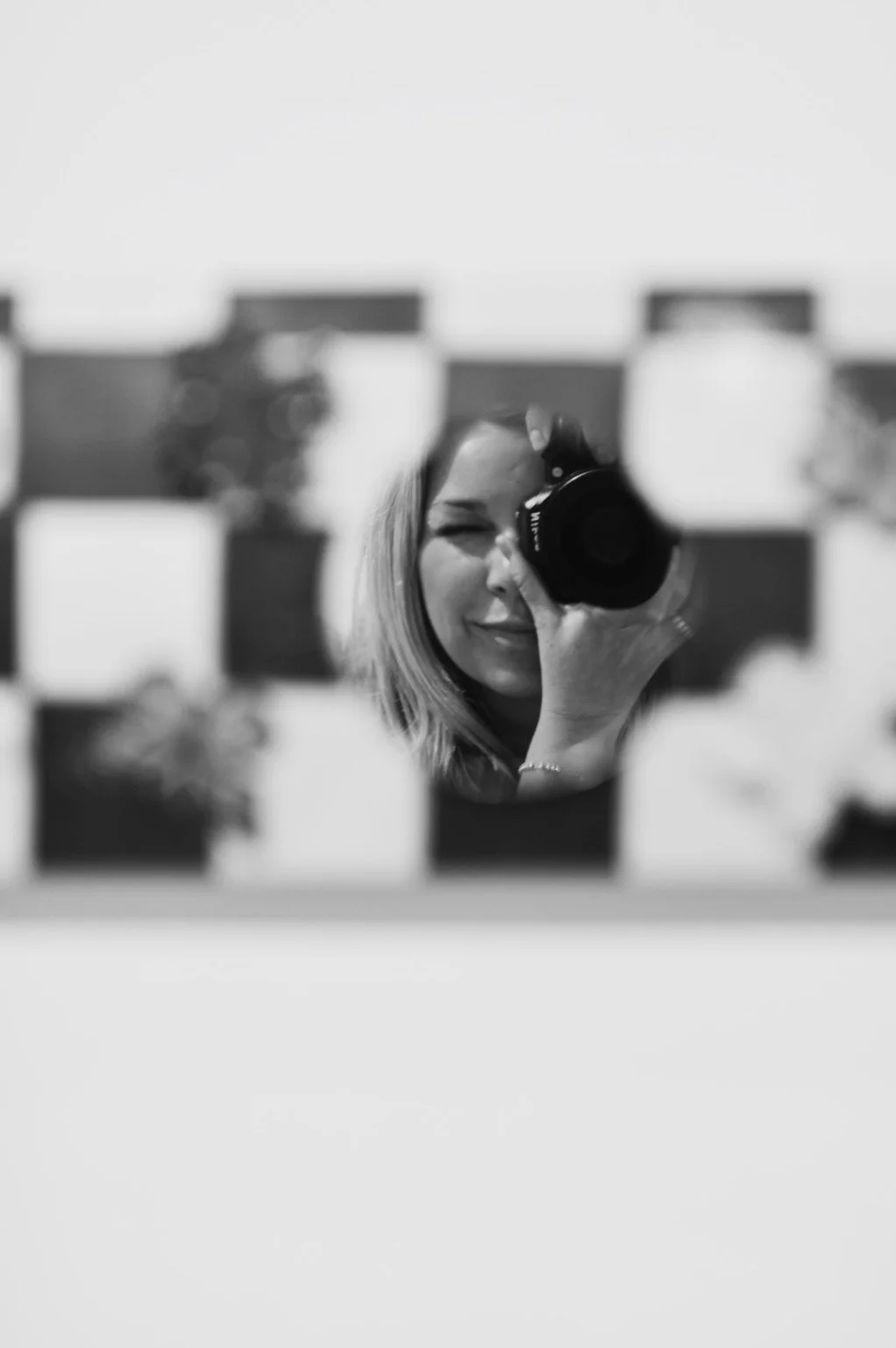 A woman taking a selfie with a camera in a mirror, with a blurred grid of small squares in the foreground.