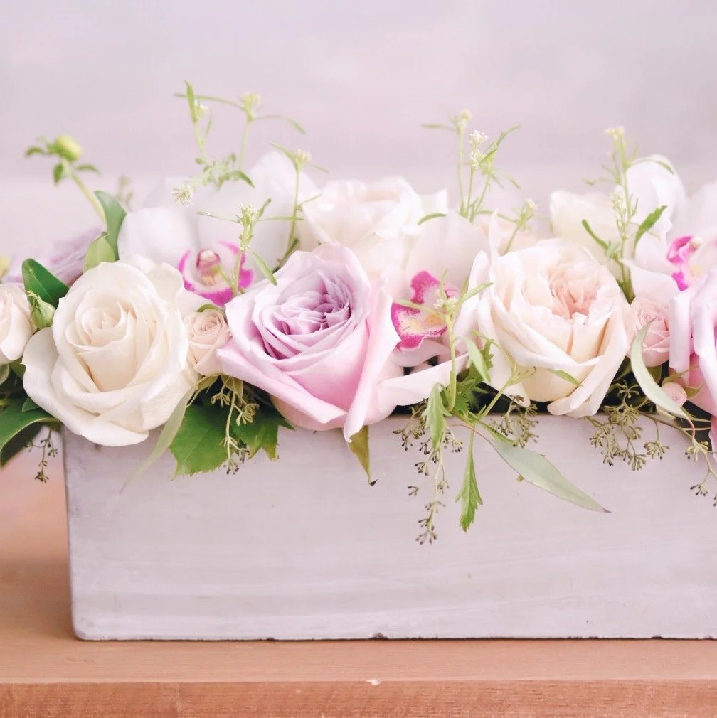 A white wooden box filled with pink and white roses and green foliage, placed on a wooden surface.