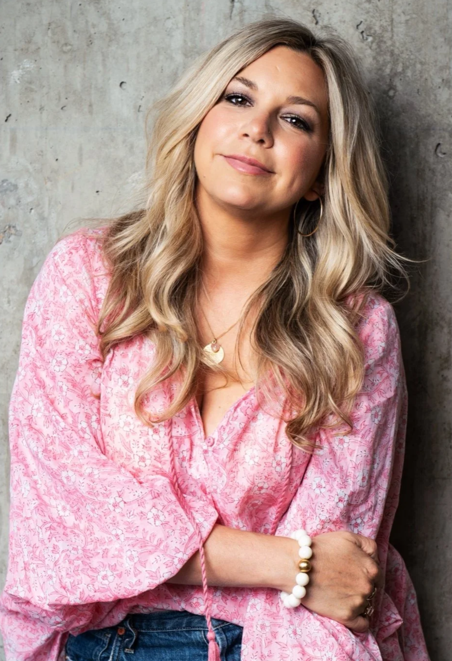 A woman with long, wavy blonde hair wearing a pink patterned blouse, blue jeans, and jewelry, standing against a concrete wall.