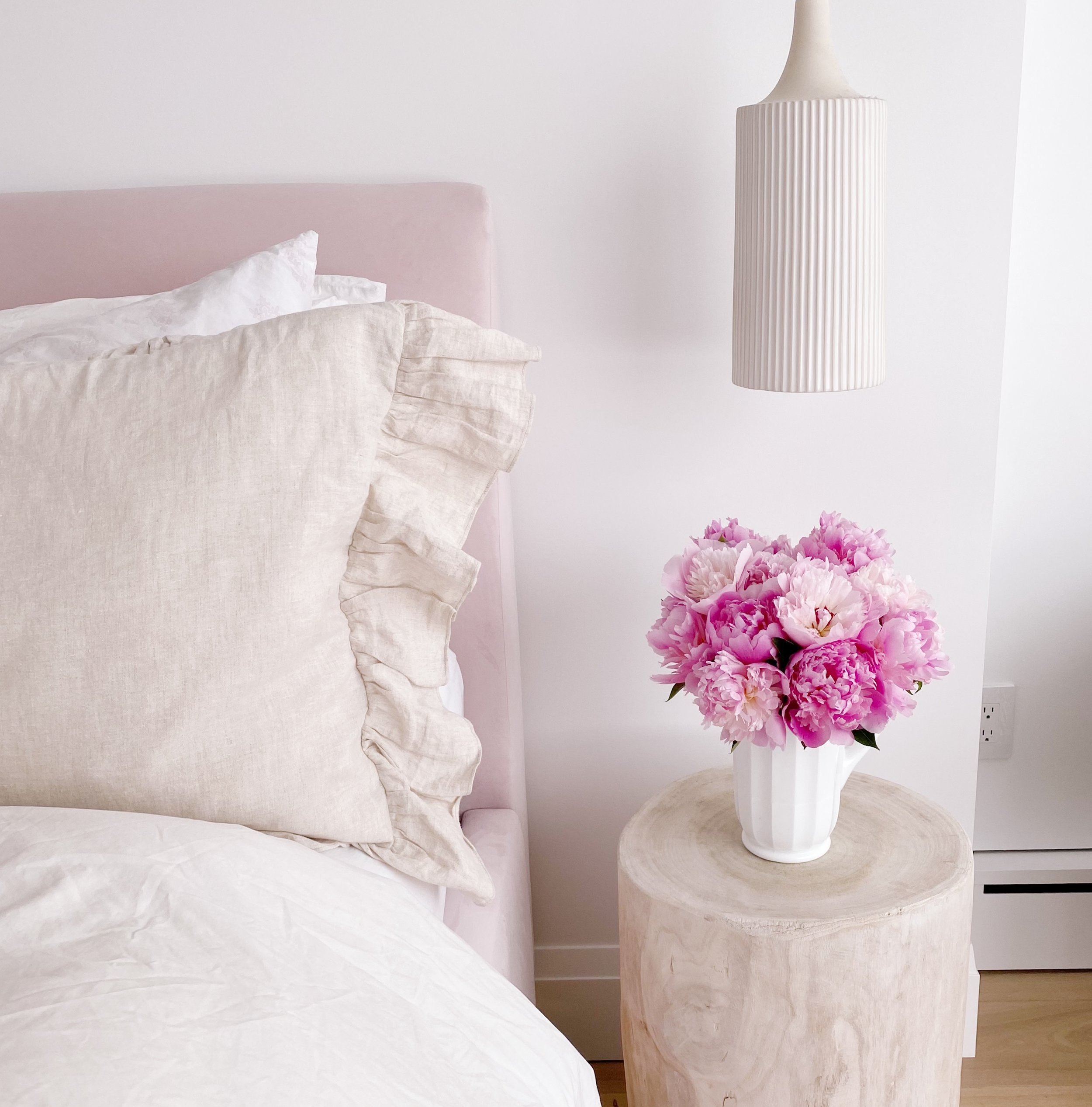 A bedroom with a bed featuring ruffled beige bedding, a pink headboard, and white pillows, a pink window frame, a white striped hanging lamp, a white vase with pink peonies on a wooden nightstand, and a white wall with an electrical outlet.
