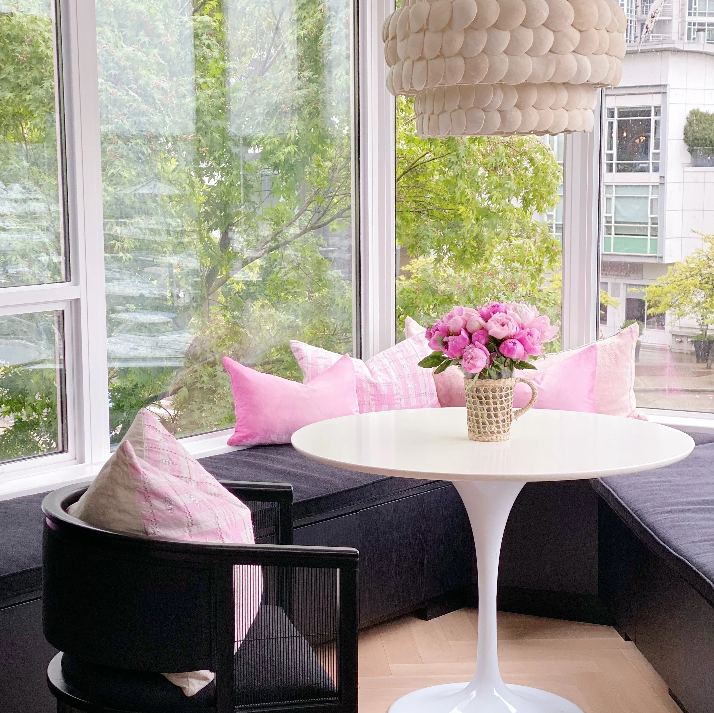 A white round table with a woven basket of pink peonies on top. A black chair with pink and white pillows is next to the table, inside a room with large windows and a view of trees and buildings outside. A beige cylindrical chandelier hangs above the