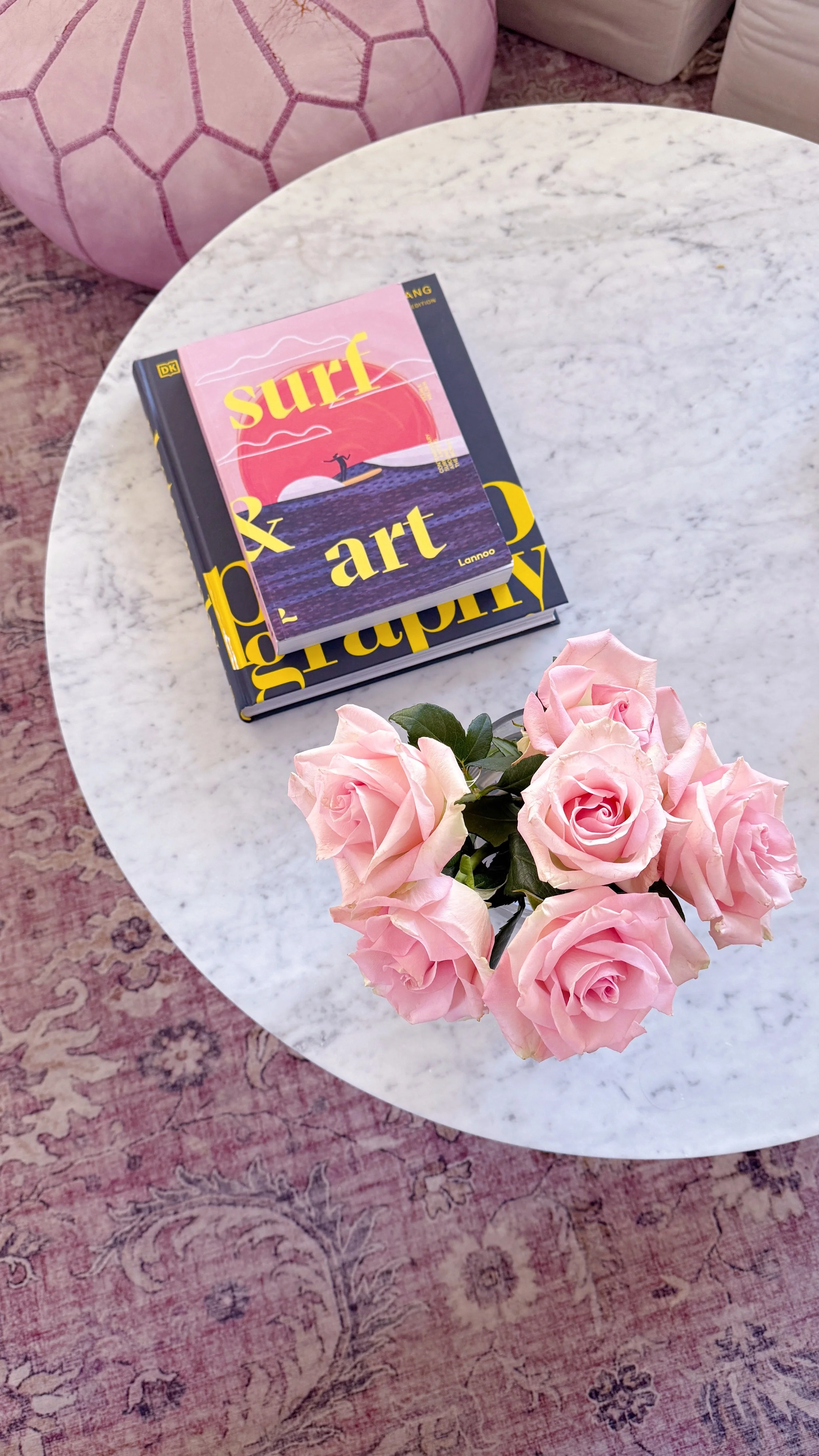 A white marble table with a bouquet of pink roses, two books stacked, and a pink round pillow with a geometric pattern in the background.