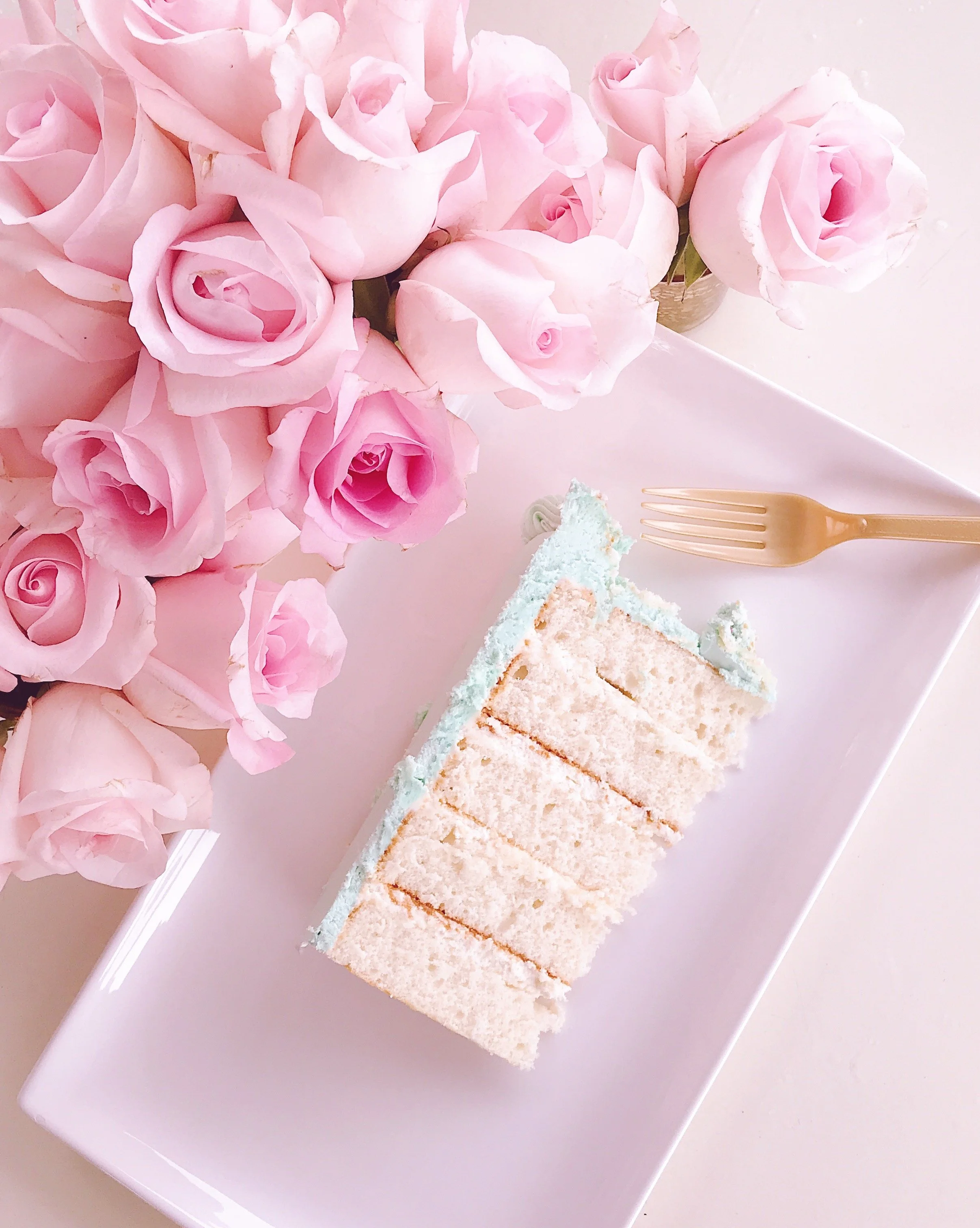 A white rectangular plate holding slices of pastel-colored cake with white frosting, next to a bunch of pink roses on a pink surface, with a gold fork to the right of the cake.