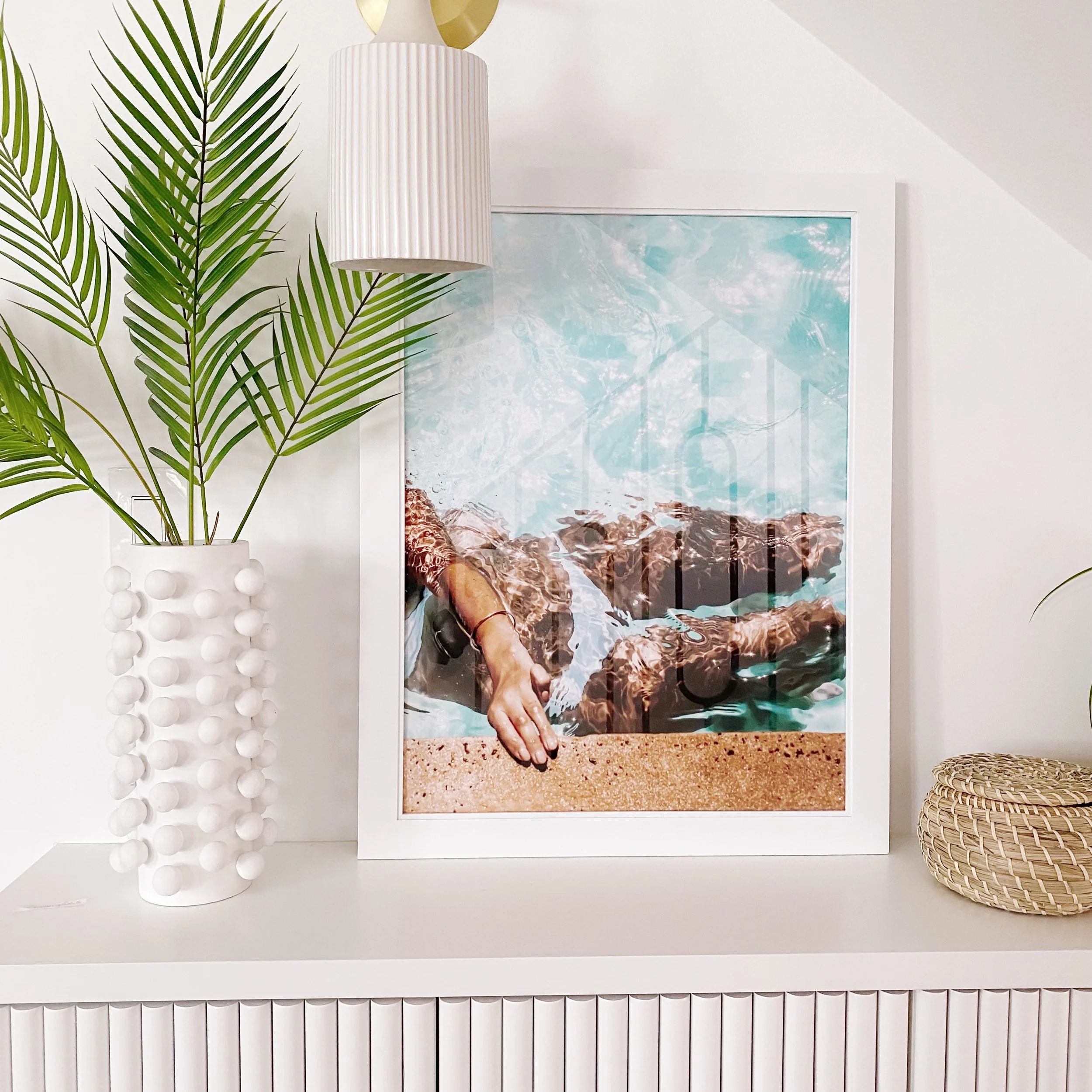 A framed beach scene with water and rocks, next to a potted palm plant, with white textured wall and decorative objects in the background.