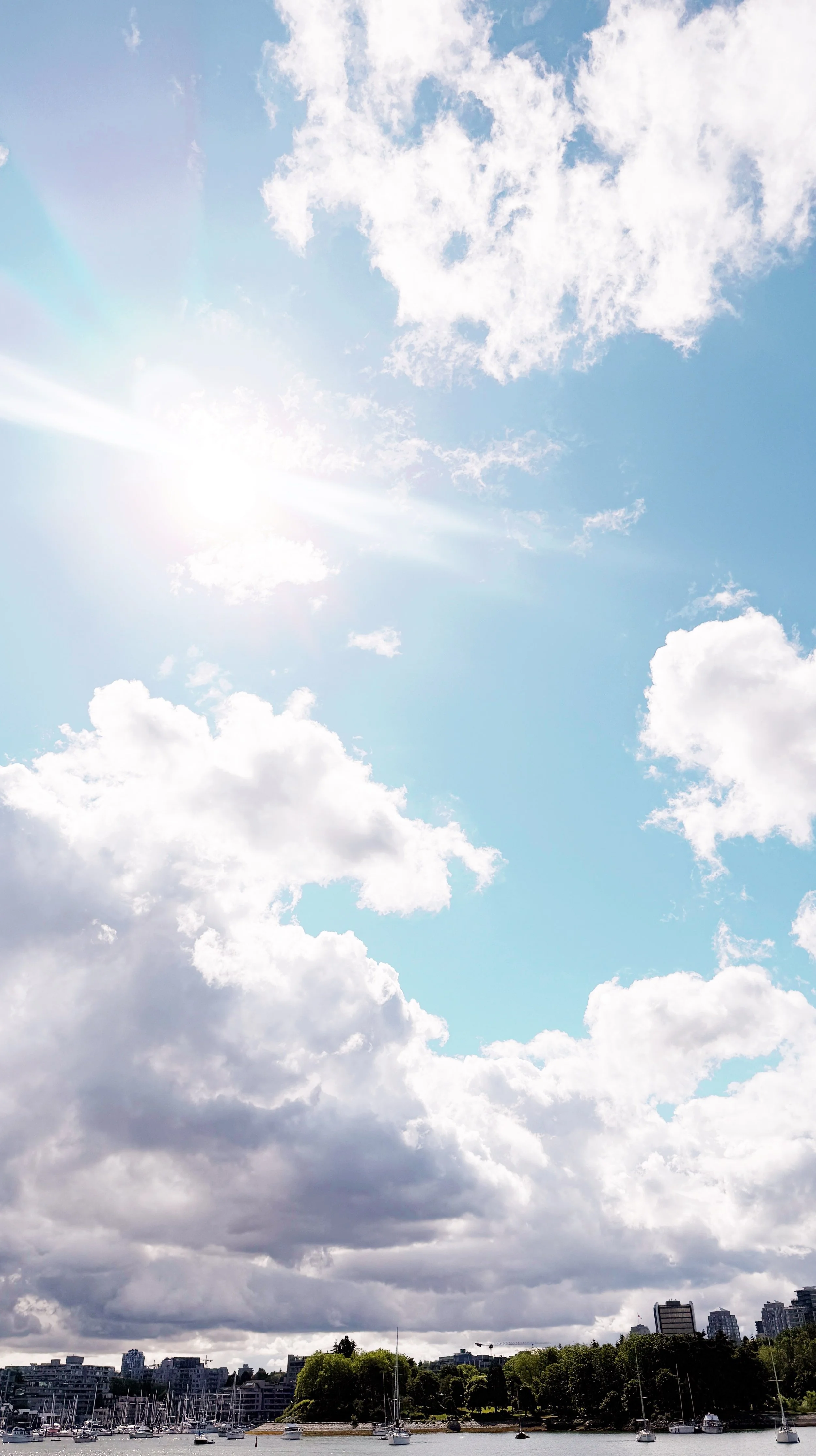 View of partly cloudy sky with the sun shining over a marina and city skyline.