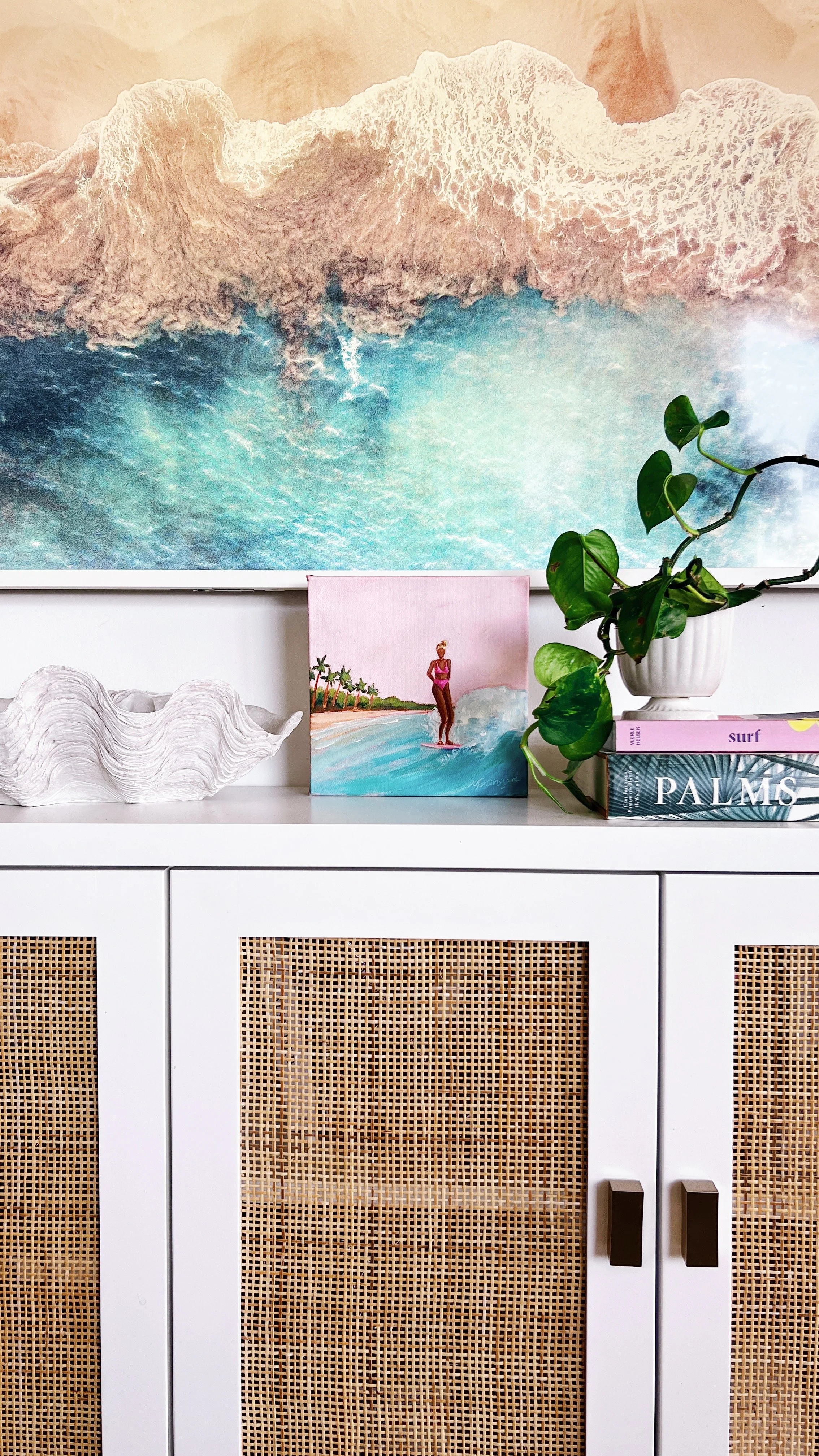 Family room or living room with a white cabinet featuring rattan panels, a large ocean-themed wall art above, a potted plant, a small portrait of a woman surfing, and books about surf and palms.