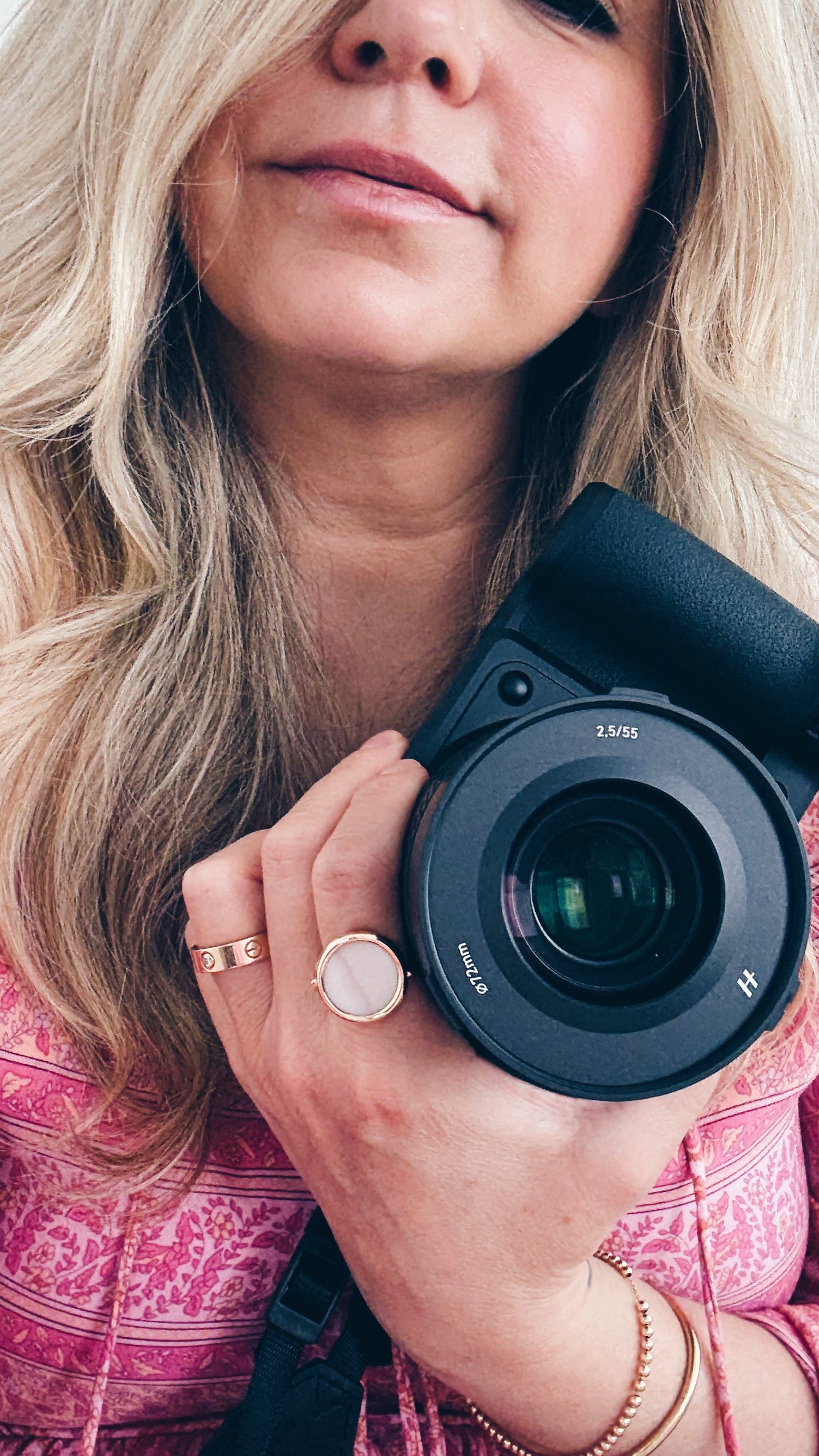 A woman with blonde hair holding a black camera and taking a selfie. She is wearing rings and a bracelet, and has on a pink patterned top.
