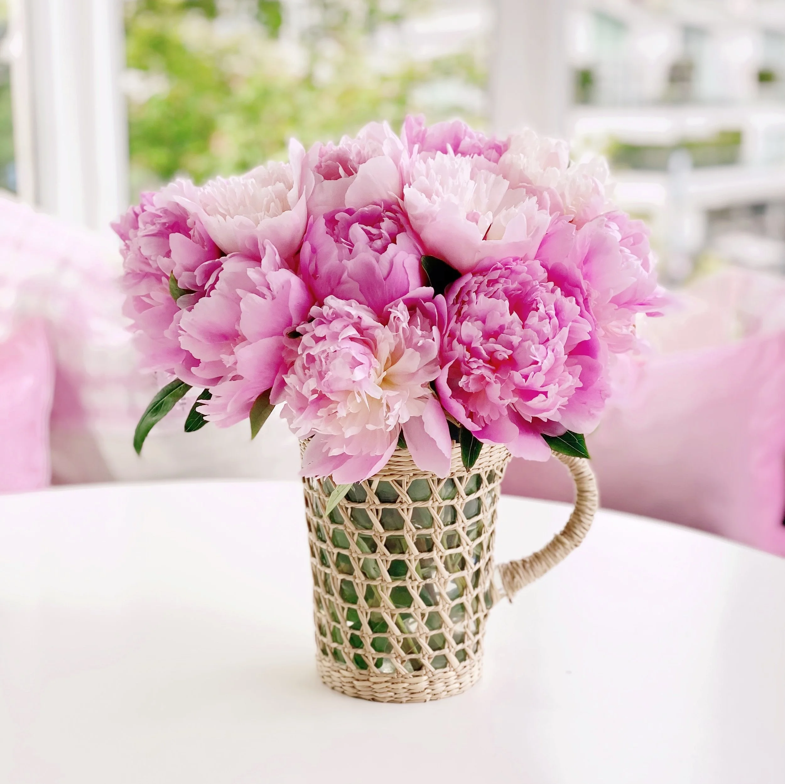 A bouquet of pink peonies in a woven basket, placed on a white surface in front of a window with a blurry green outdoor background.