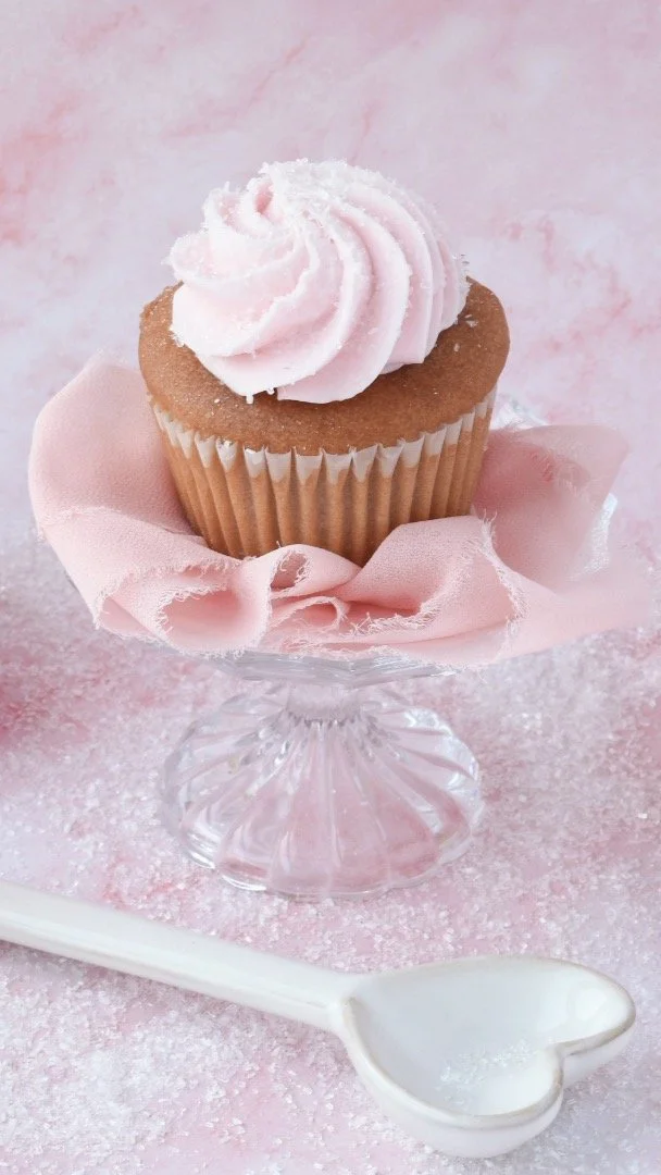 A cupcake with pink frosting on a glass cake stand, surrounded by pink fabric and a pink background.