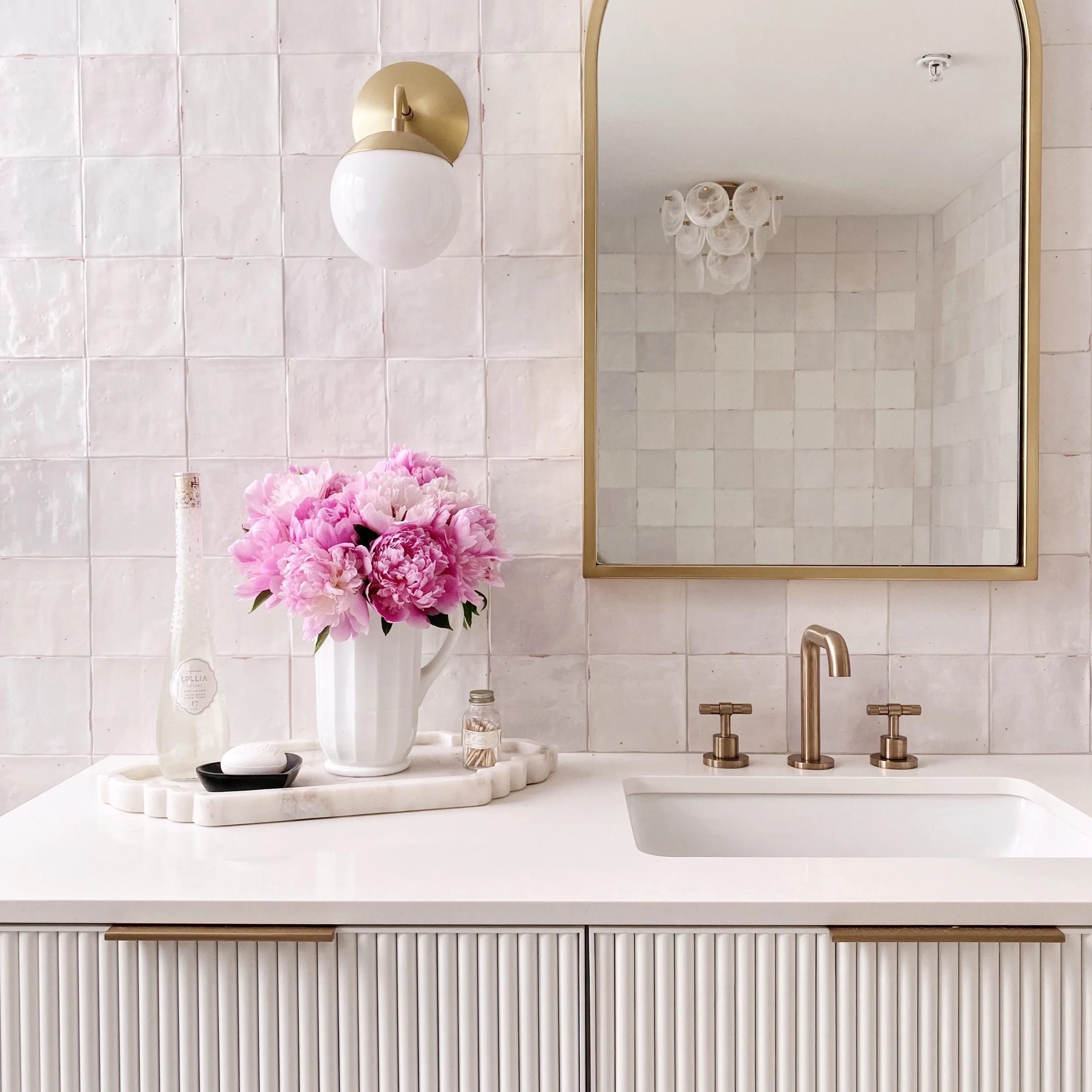 A bathroom mirror above a white marble countertop with a vase of pink flowers, a soap dispenser, and a small container. The background features white tiled walls and a brass wall sconce with a white glass globe. The mirror reflects a white ceiling wi