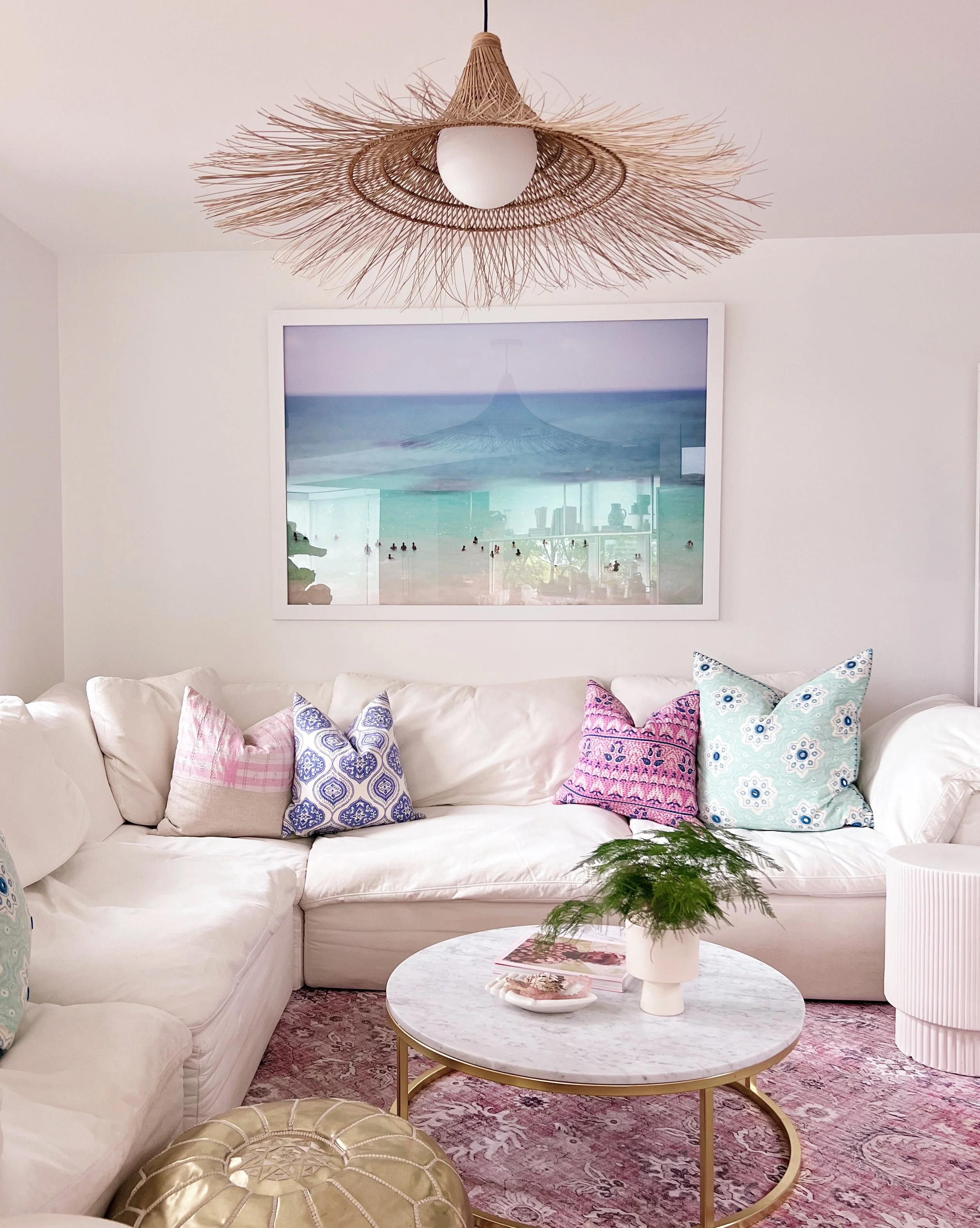 Living room with a white sofa, colorful patterned throw pillows, a marble top coffee table with a magazine and a white vase with green fern, framed beach scene picture on the wall, and a woven pendant light fixture hanging from the ceiling.