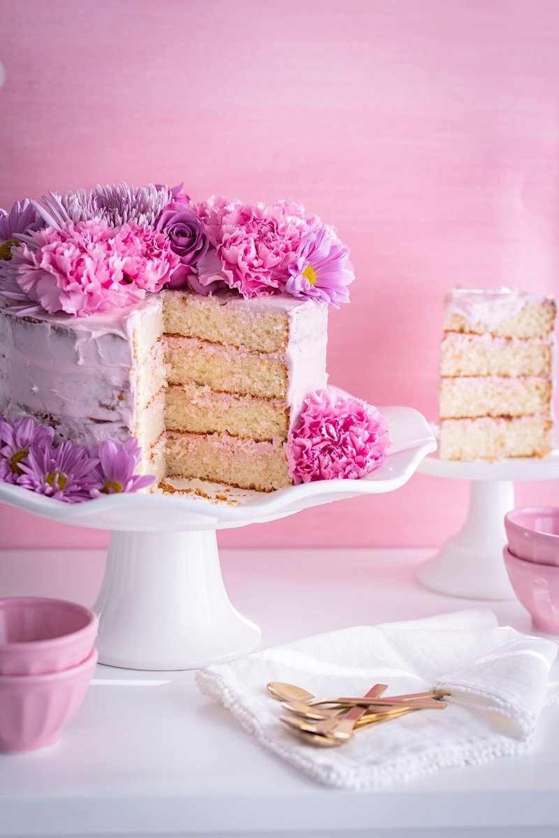 A pink floral layered cake with white icing on a white cake stand, decorated with pink and purple flowers, with a slice cut out. There are pink bowls, gold utensils, and a white cloth in front of the cake against a pink background.